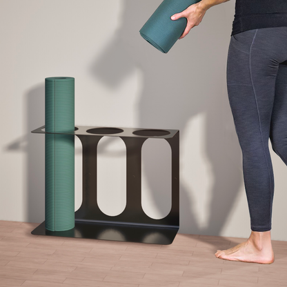 Black metal yoga mat wall bracket holding a green rolled yoga mat while a person places another rolled yoga mat beside it. — for Carpe Diem Dream