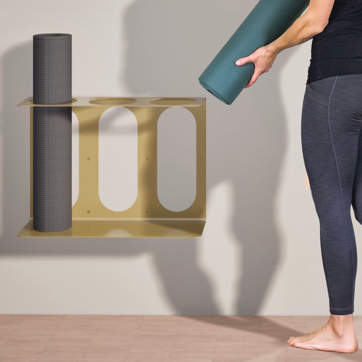 Gold metal yoga mat wall bracket holding a gray rolled yoga mat while a person places a green rolled yoga mat beside it. — for Carpe Diem Dream